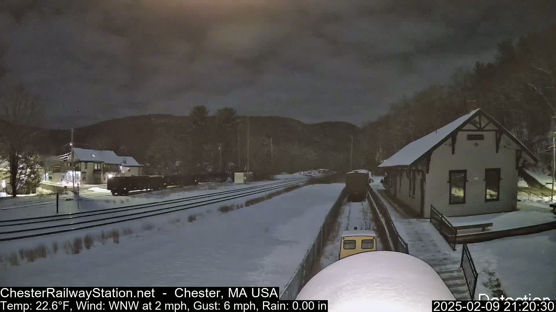 A snow-covered train station and railroad tracks are visible at night under a dark, cloudy sky, with a train partially visible.