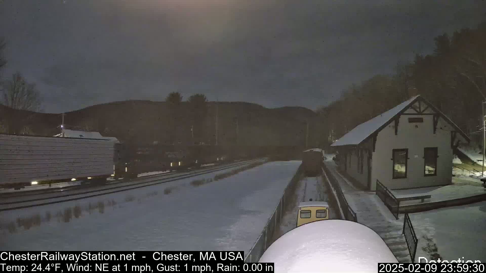 A snow-covered train station and railway yard are visible at night under a dark sky, with a train partially visible in the distance.