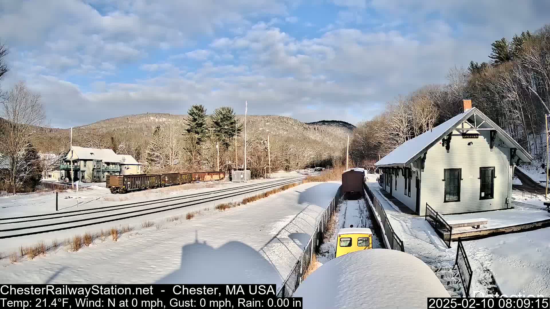 A snow-covered train station and railway tracks are seen, with a small yellow train approaching and freight cars in the background under a partly sunny sky.
