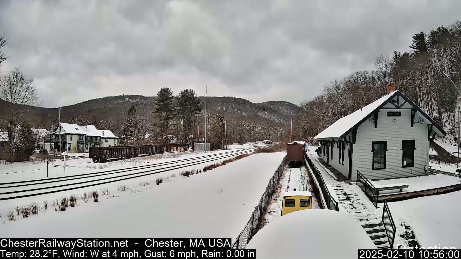 A snow-covered train station and railway tracks are seen under an overcast sky, with a small yellow train car and freight cars present.