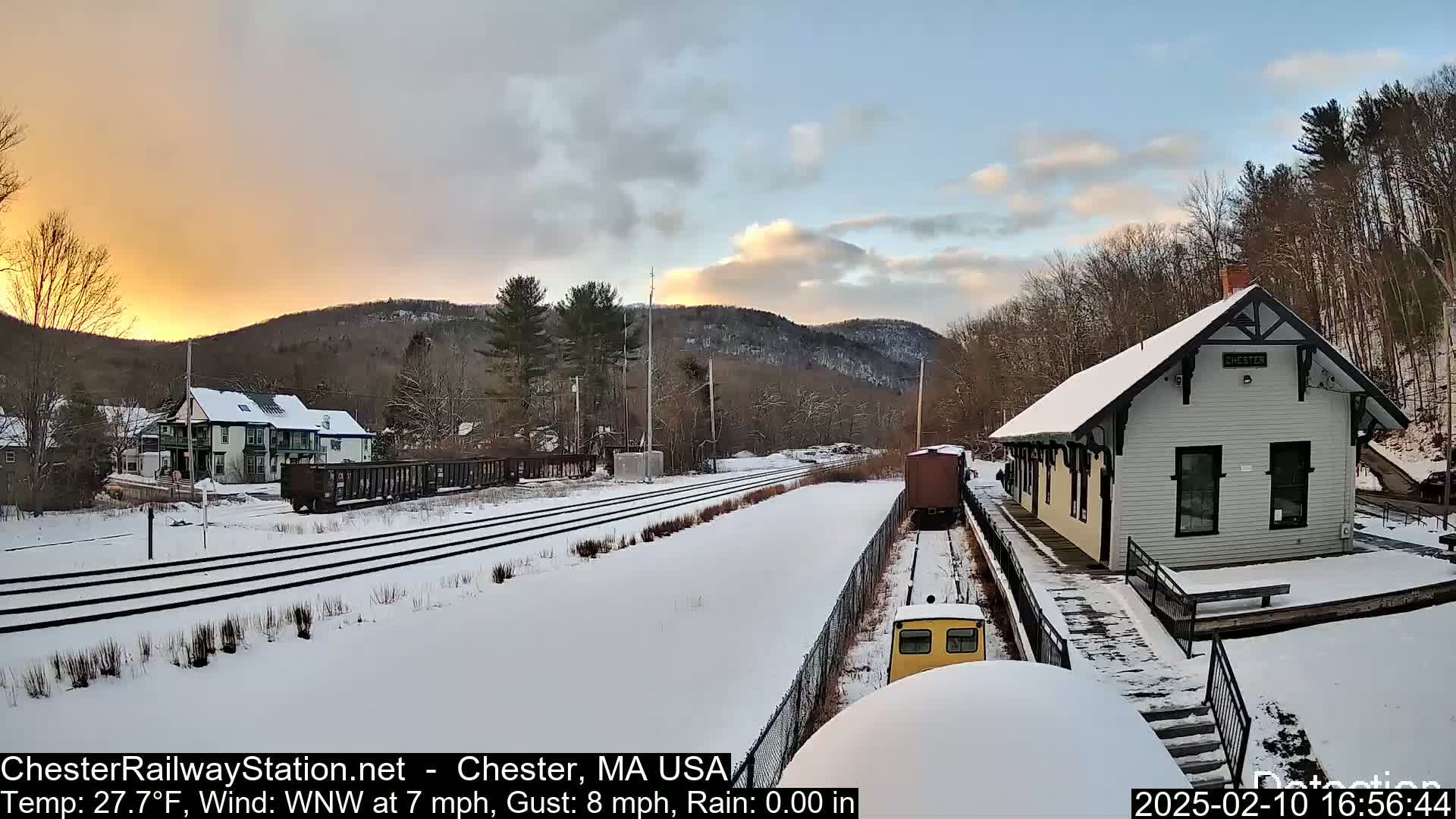 A snow-covered train station and tracks are visible at sunset, with a train car nearby, and mountains in the background under a partly cloudy sky.