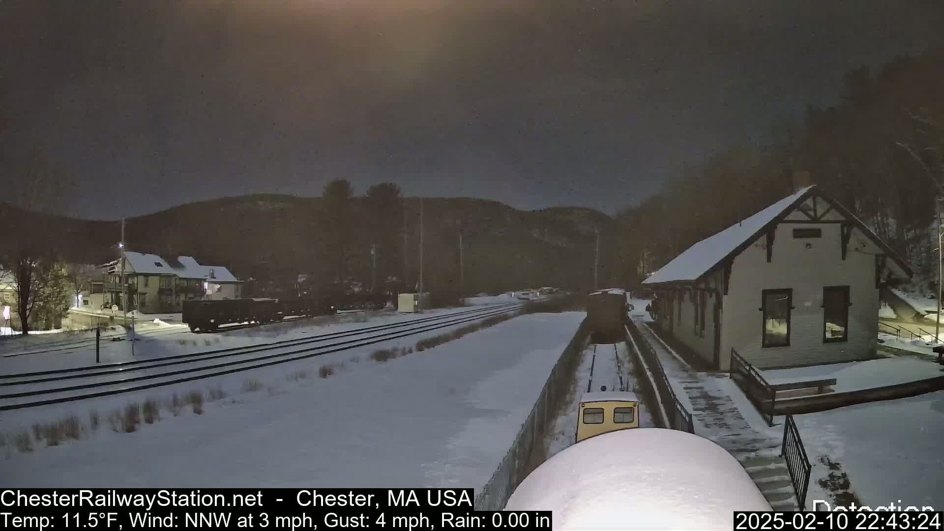 A snow-covered train station and railway tracks are visible under a dark night sky, with a train approaching the station.