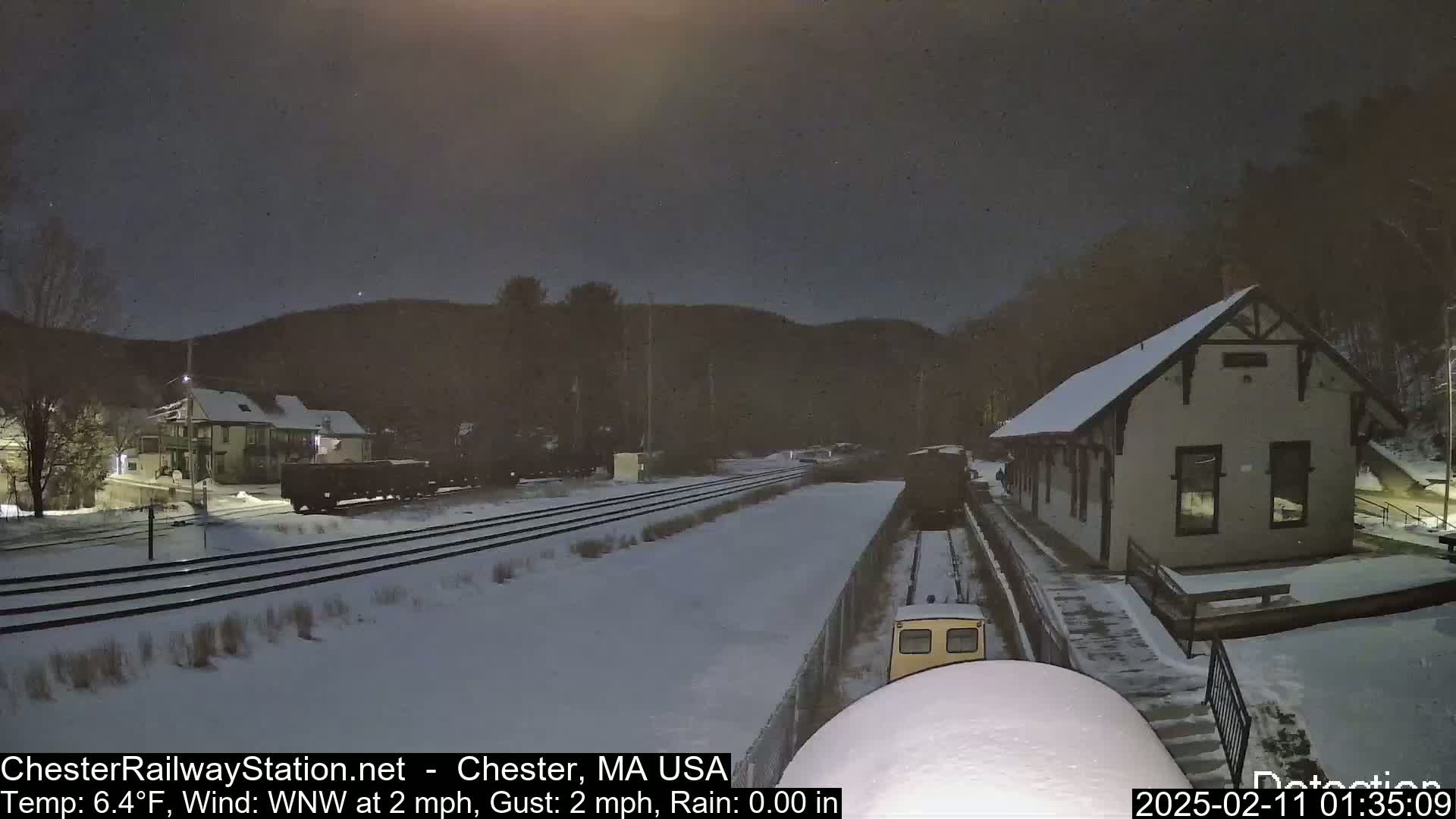 A snow-covered train station and railroad tracks are visible under a dark night sky, with a train partially obscured by snow in the foreground.