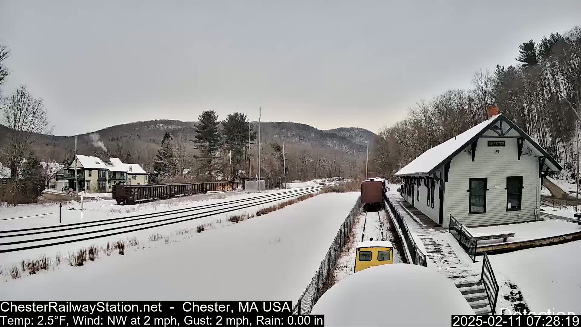 A snow-covered train station sits beside railroad tracks with several train cars and a small yellow vehicle, all against a backdrop of snow-covered hills and trees under an overcast sky.