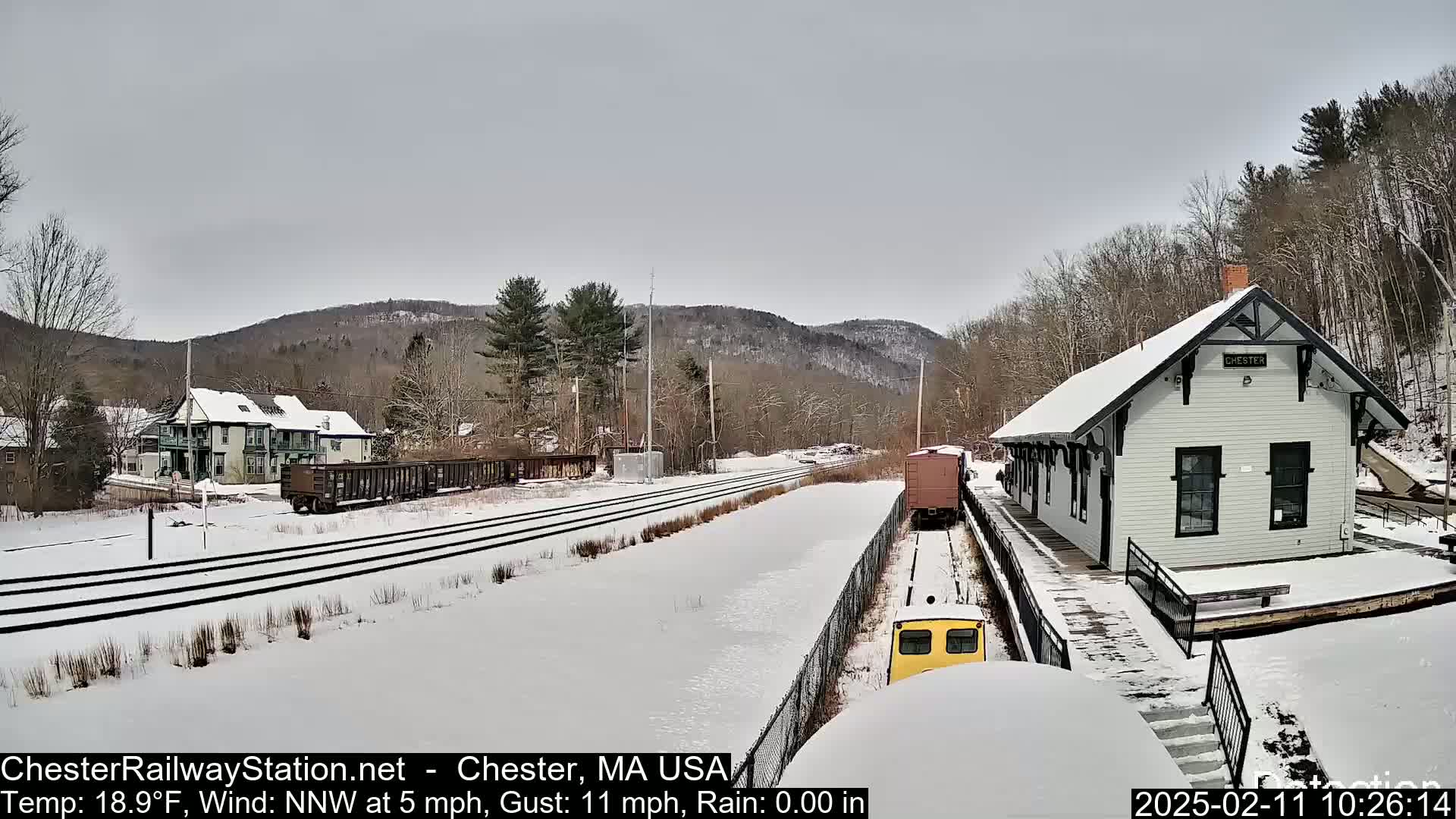 A snow-covered train station and rail yard are situated in a valley, with a building and rail cars visible under a cloudy sky.