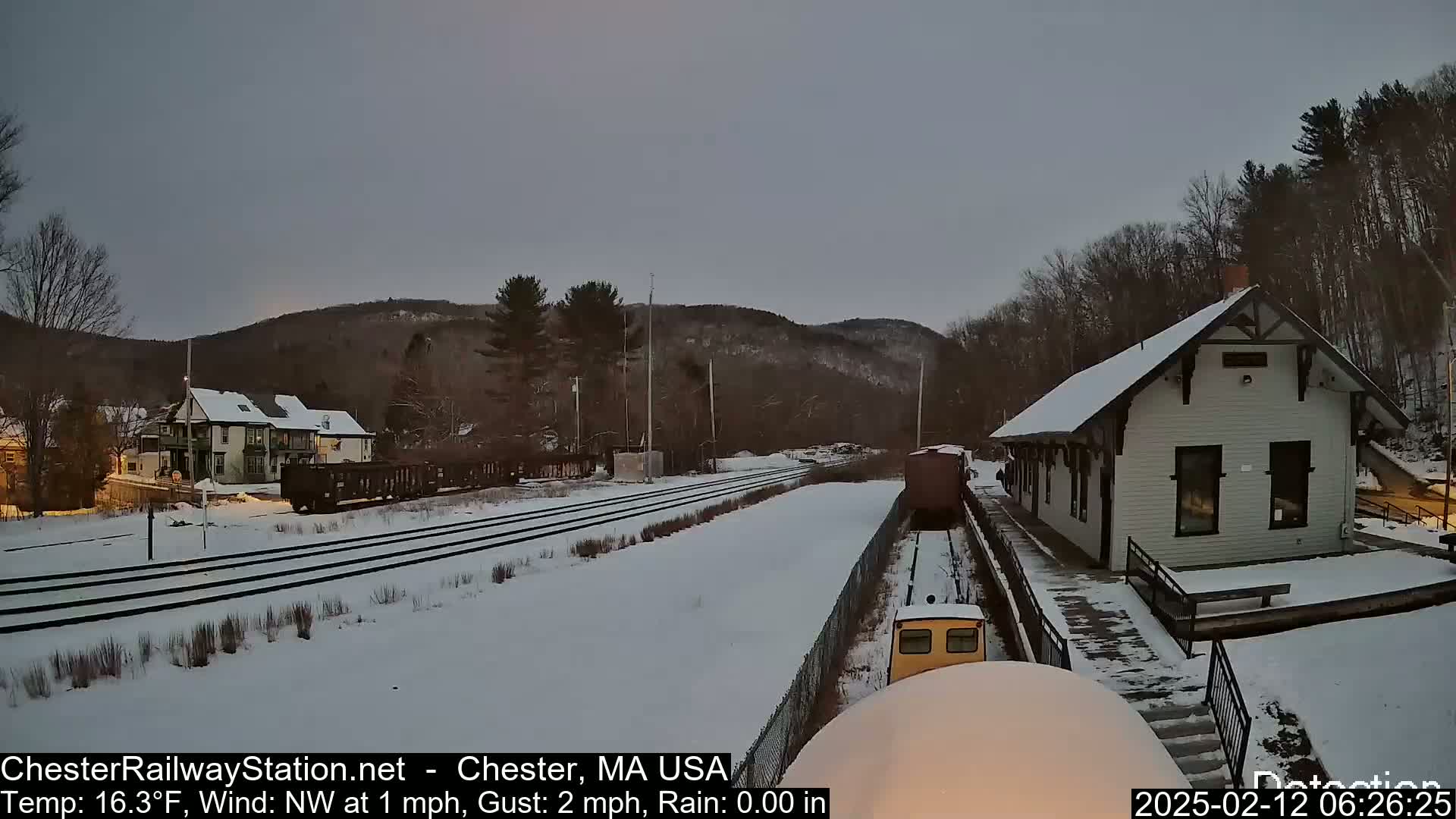 A small yellow train approaches a snow-covered train station next to a snowy landscape under an overcast sky.