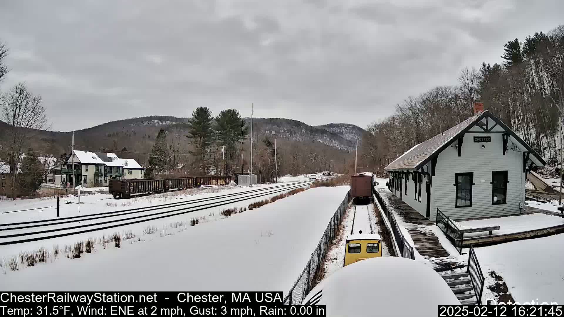 A snow-covered train station sits beside a railway with a small train approaching, nestled among snowy hills under a cloudy sky.