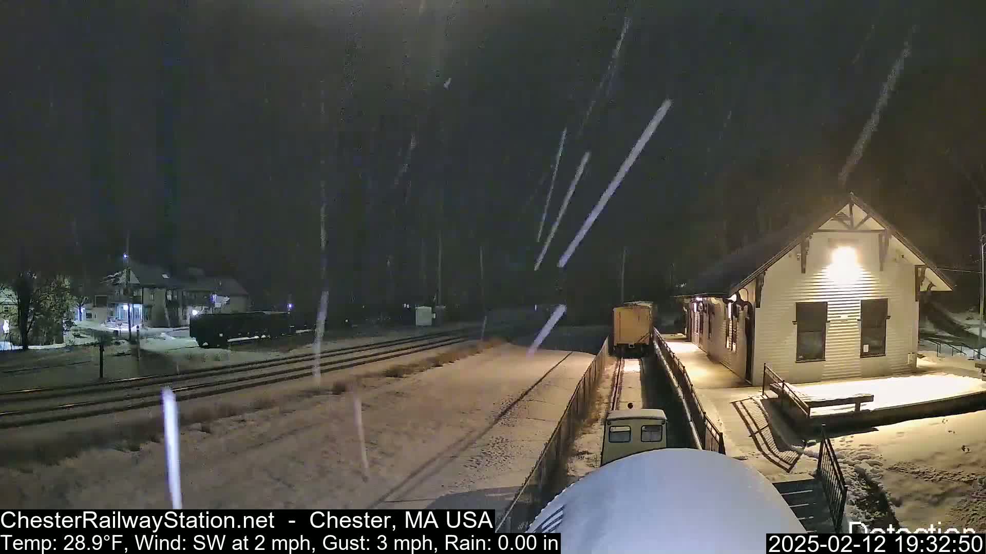 A snow-covered train station and railway tracks are seen at night during a light snowfall.