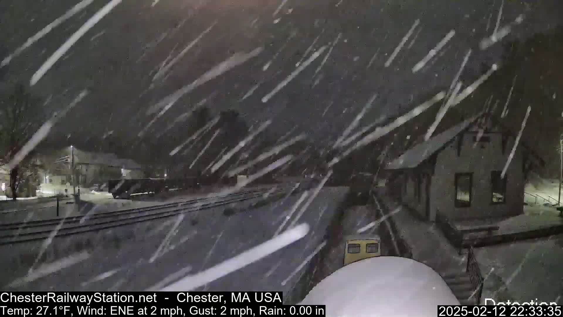 Heavy snow falls on a train at a station near a small building.