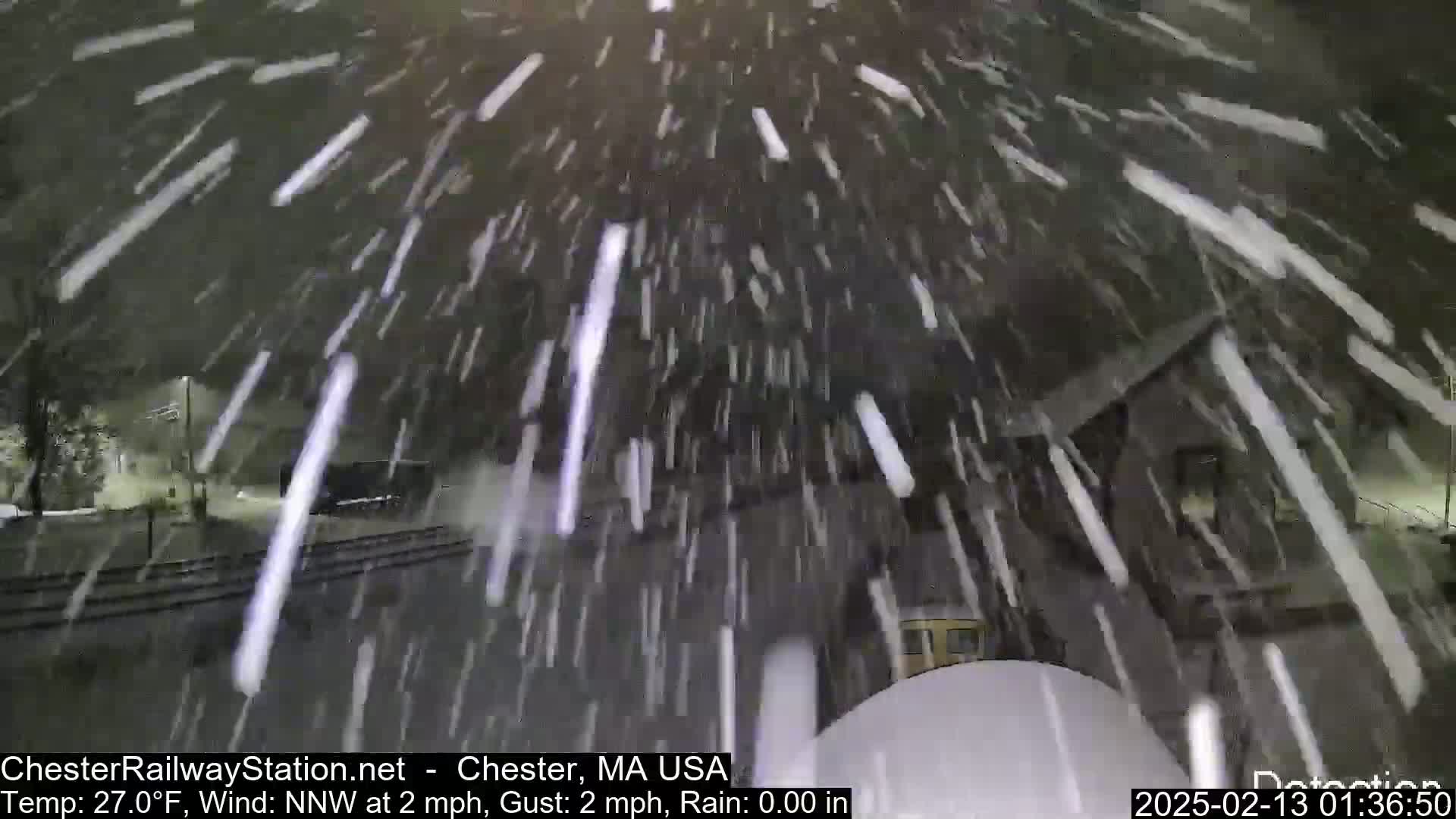 Heavy snow falls at night near a train track and building.