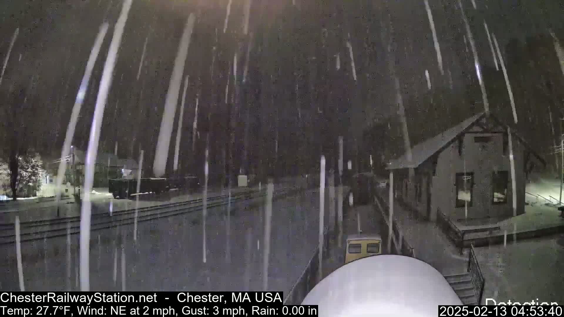 A nighttime scene of snow falling heavily over a train station and railway tracks.
