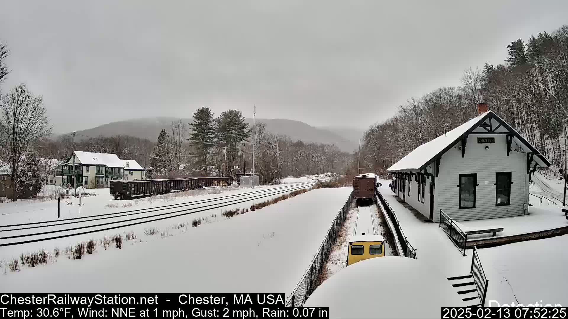 A snow-covered train station sits beside a railway track, with freight cars and a small building visible, under an overcast sky with light rain.