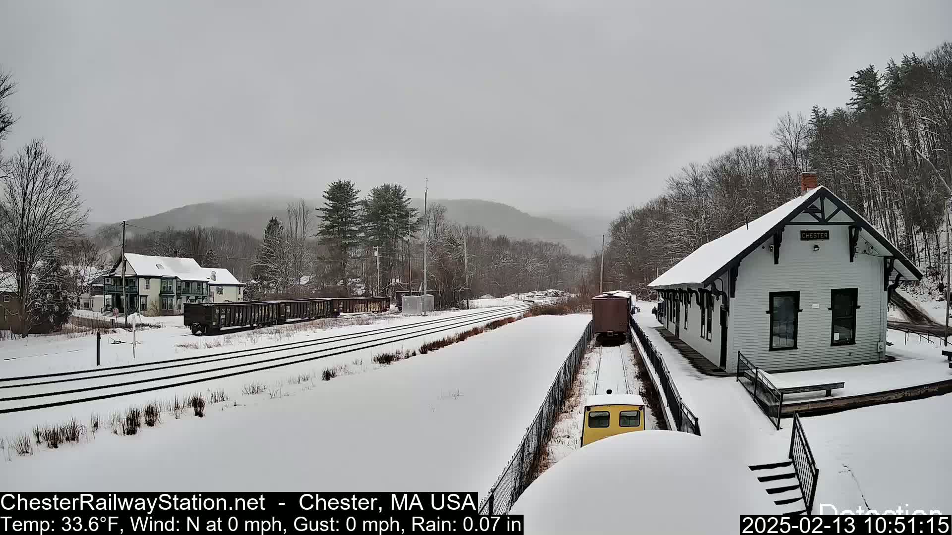 A snow-covered train station and tracks are visible, with several train cars and a small train present in the scene, under an overcast sky.