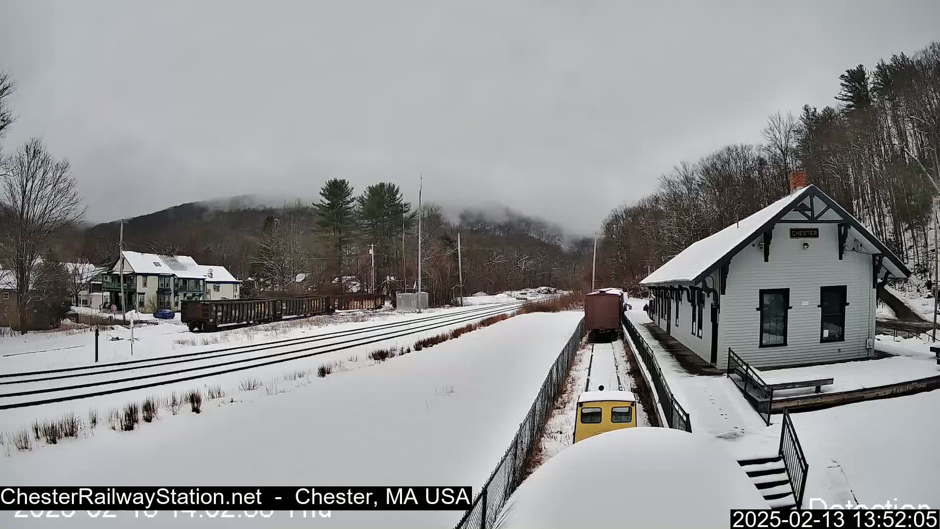 A snow-covered train station and railway lines are visible, with a train car in the foreground and a mountain shrouded in fog in the background under an overcast sky.