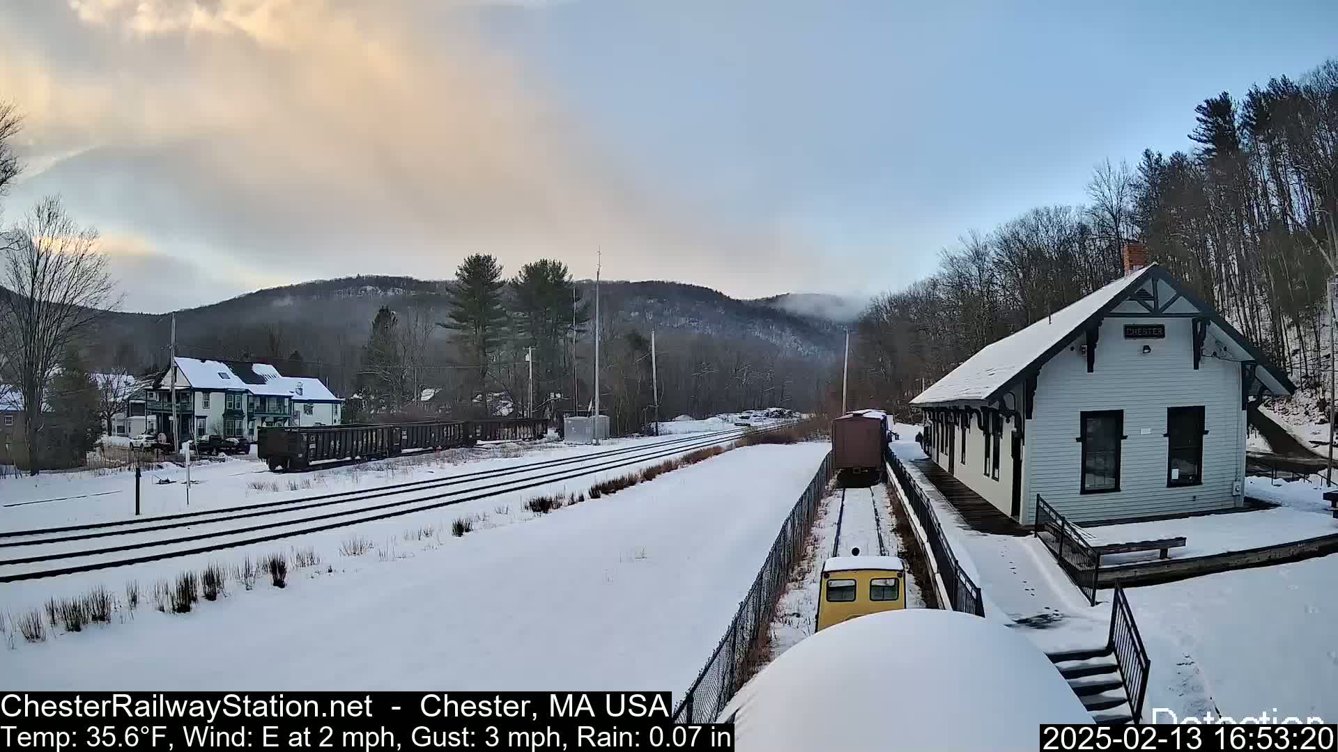 A snow-covered train station and railway tracks are visible, with a light dusting of snow on the ground and hills in the background under a partly cloudy sky.
