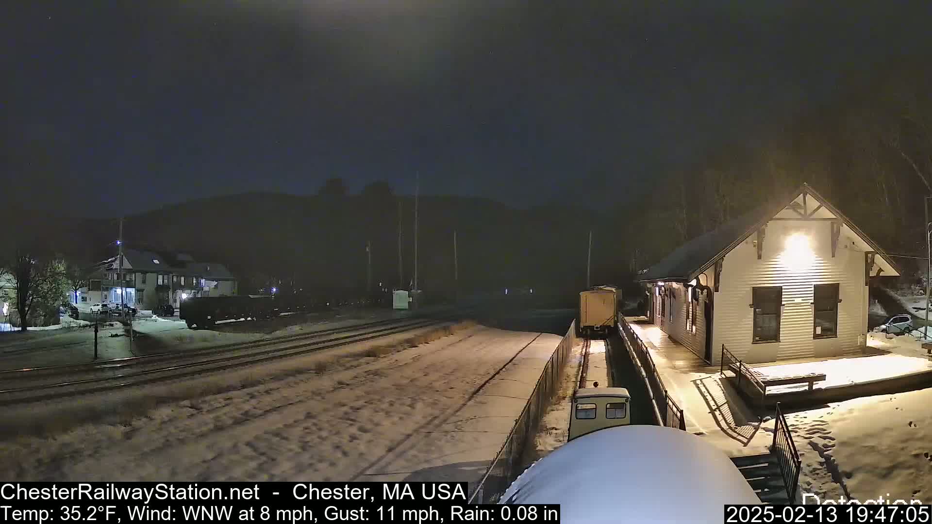 A nighttime scene of a snow-covered train station and railway with a train visible, under a dark sky with light rain.