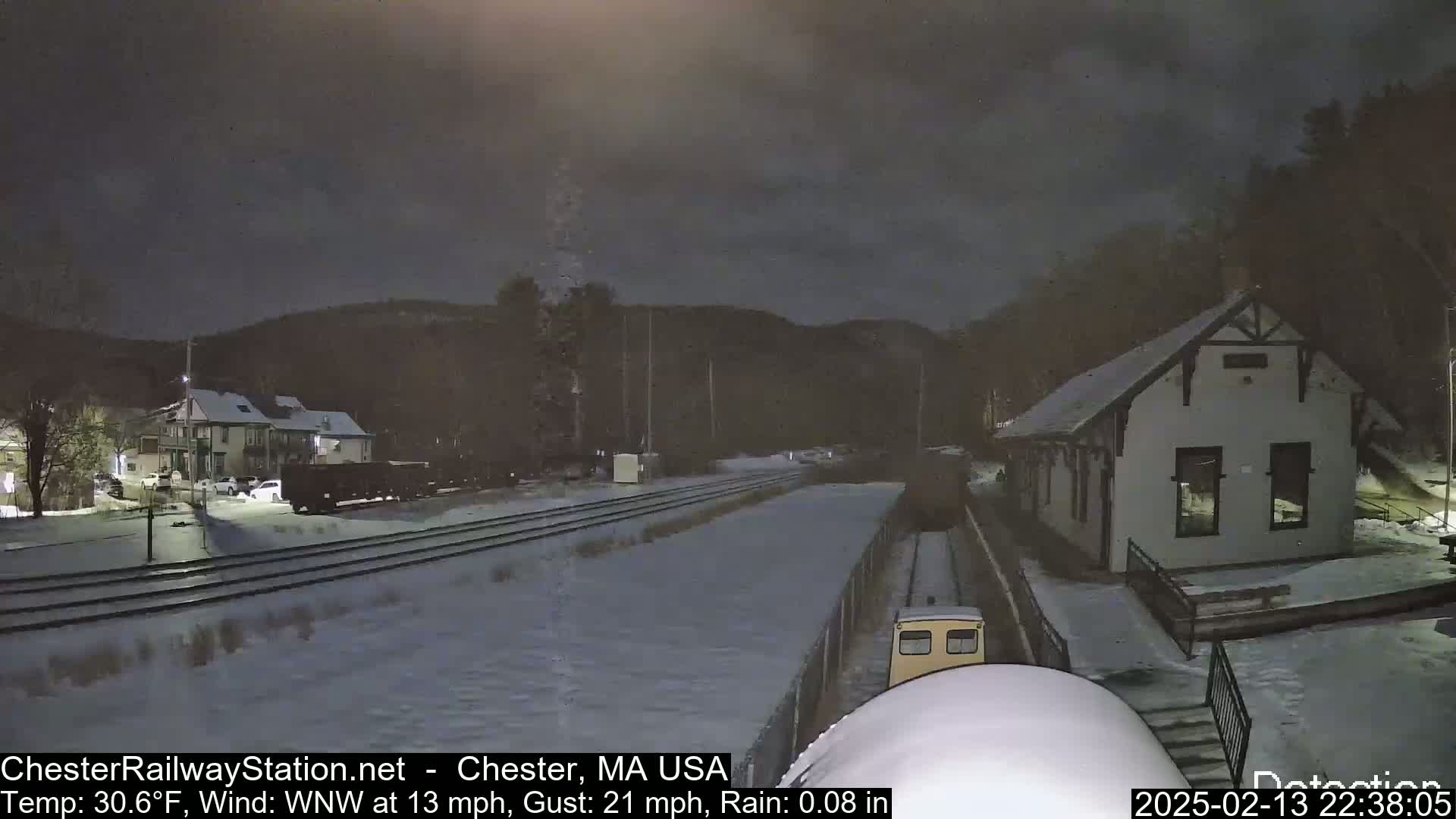 A snow-covered train yard at night with a small train approaching a station building under a dark, cloudy sky.