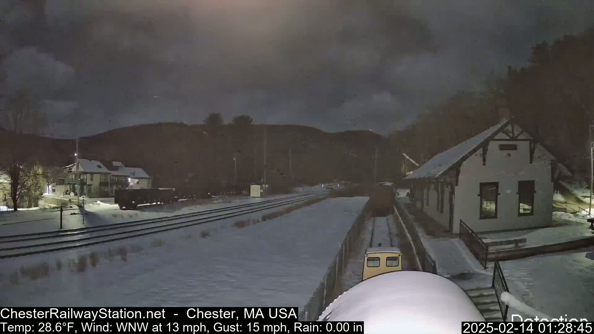 A snow-covered train yard at night, with a small building and a train visible, under a dark, cloudy sky.