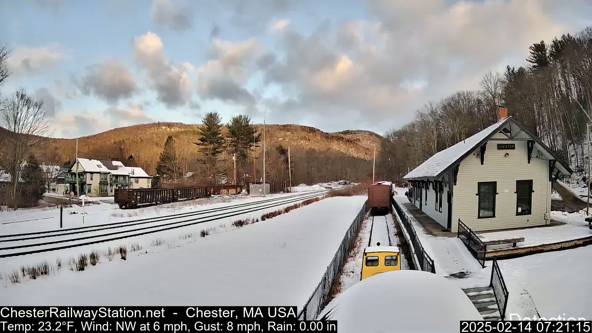 A snow-covered train station and railroad tracks are visible, with a train partially seen, under a partly cloudy sky.