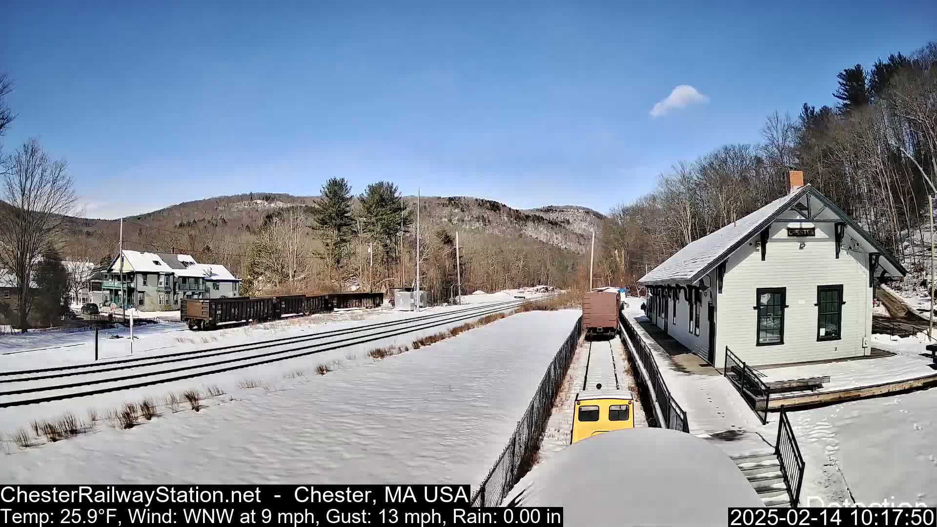 A snow-covered train station and tracks are visible under a mostly clear, sunny sky, with a few freight cars and a small yellow train car present.