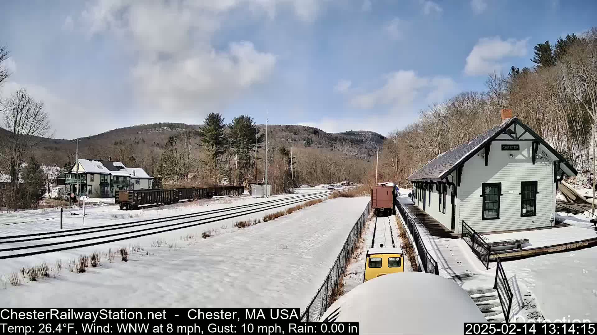 A snow-covered train station and railway tracks are seen under a partly cloudy sky, with a small train approaching the station.