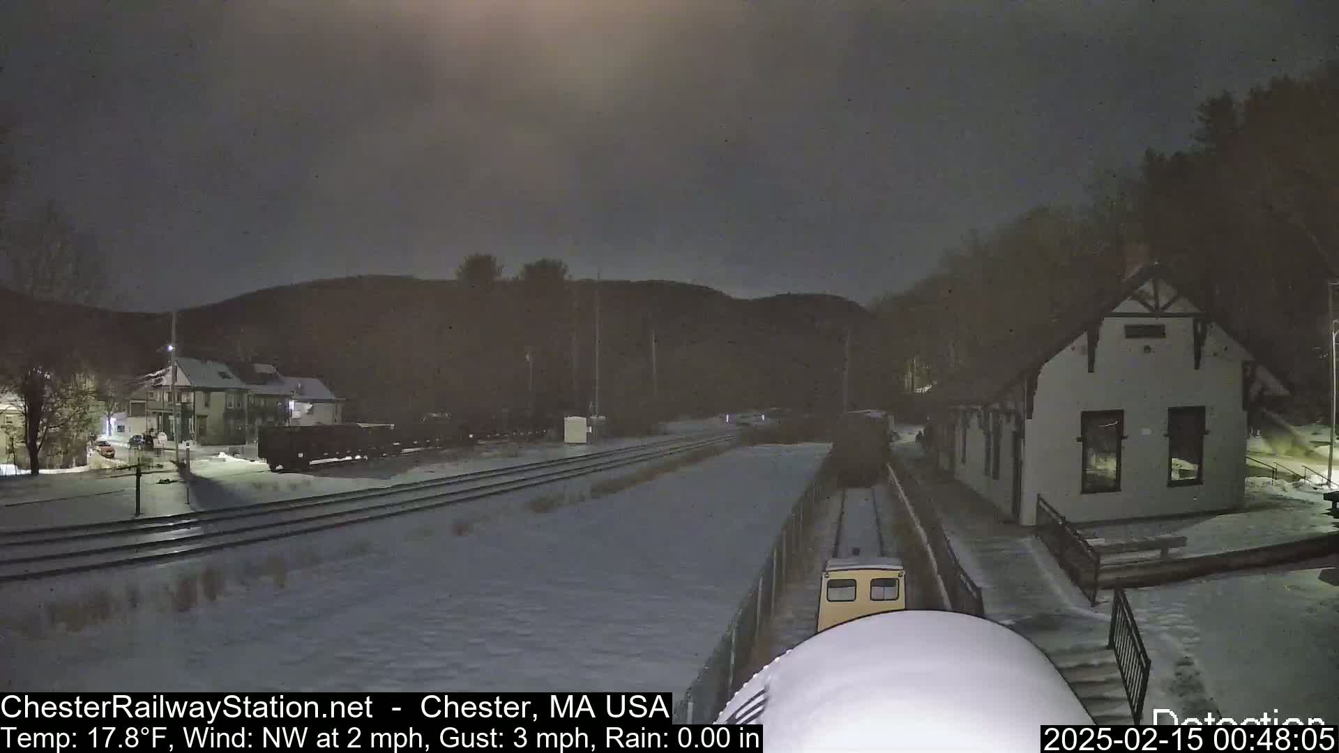 A snow-covered train approaches a small station building under a dark, nighttime sky.