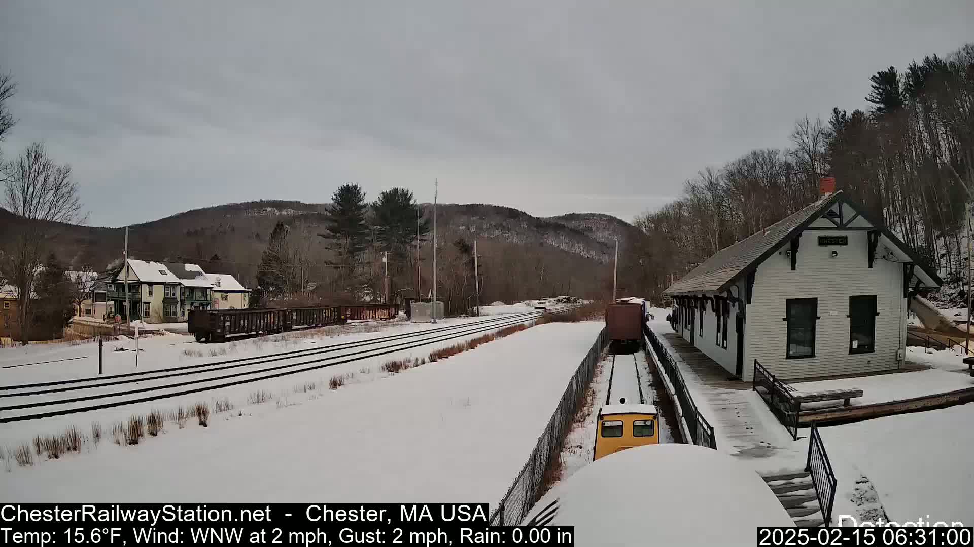A snow-covered train station and tracks are visible, with a train approaching the station under a cloudy sky.