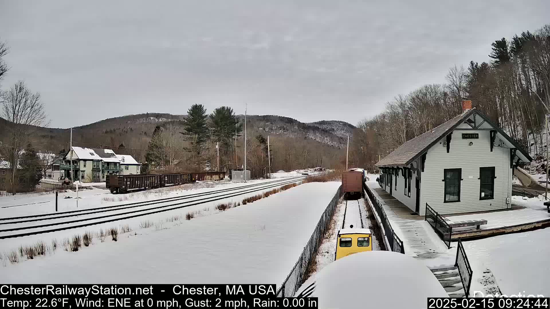A snow-covered train station and tracks are visible, with a train partially in view, under a cloudy sky.