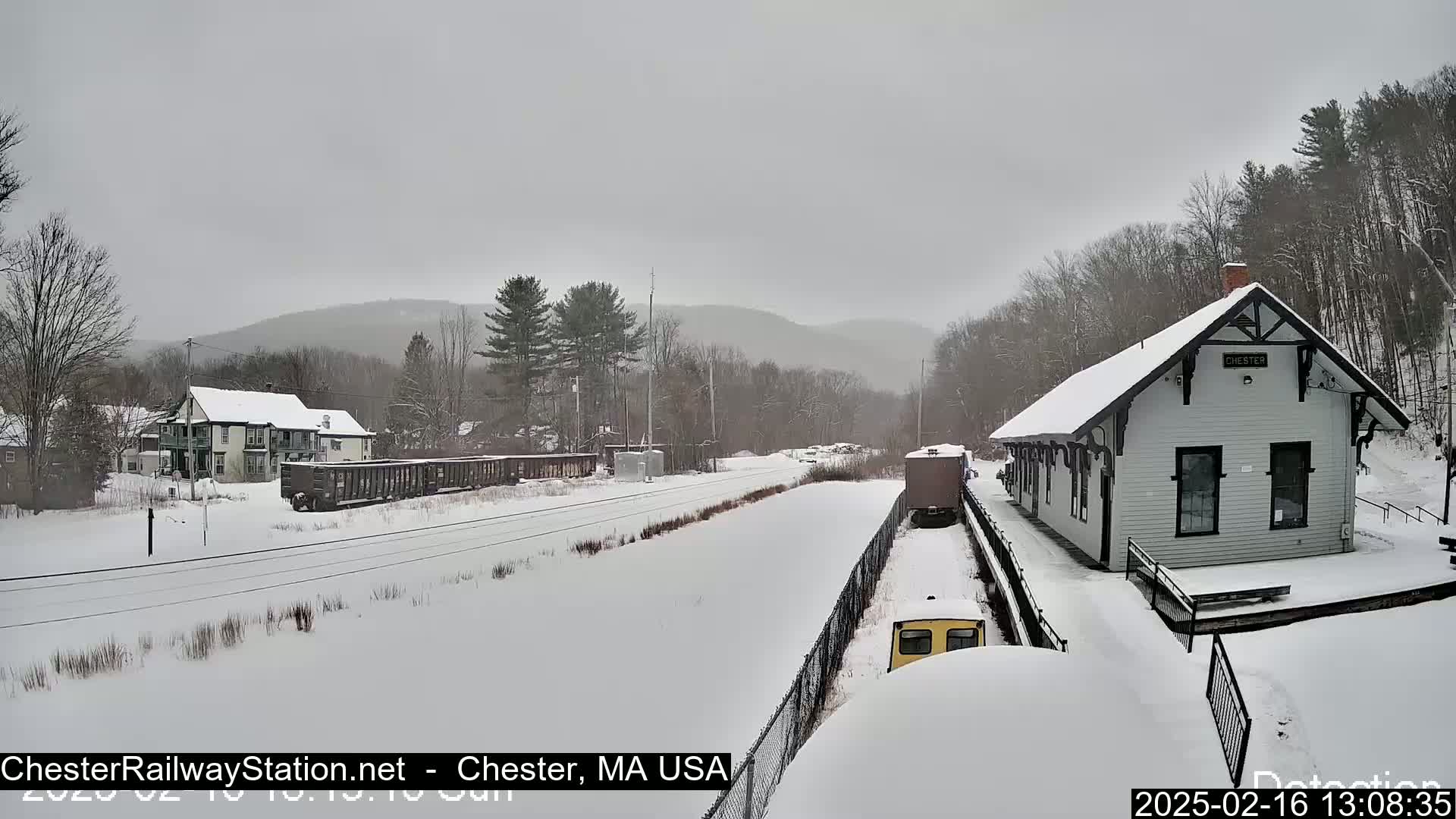 A snow-covered train station and railway line are visible, with a light snowfall and distant hills in the background.