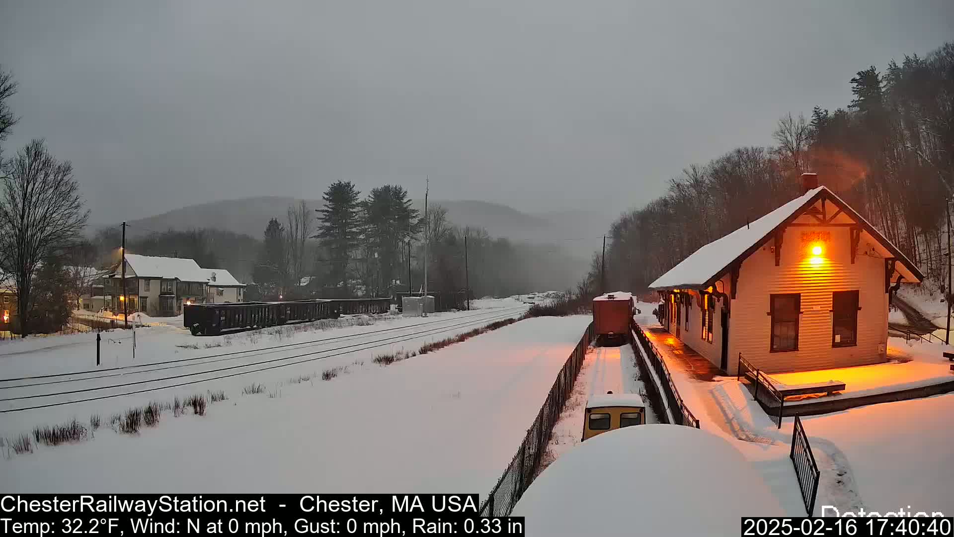 A snow-covered train station is illuminated at night, with a train car visible in the distance and light rain falling under a gray sky.