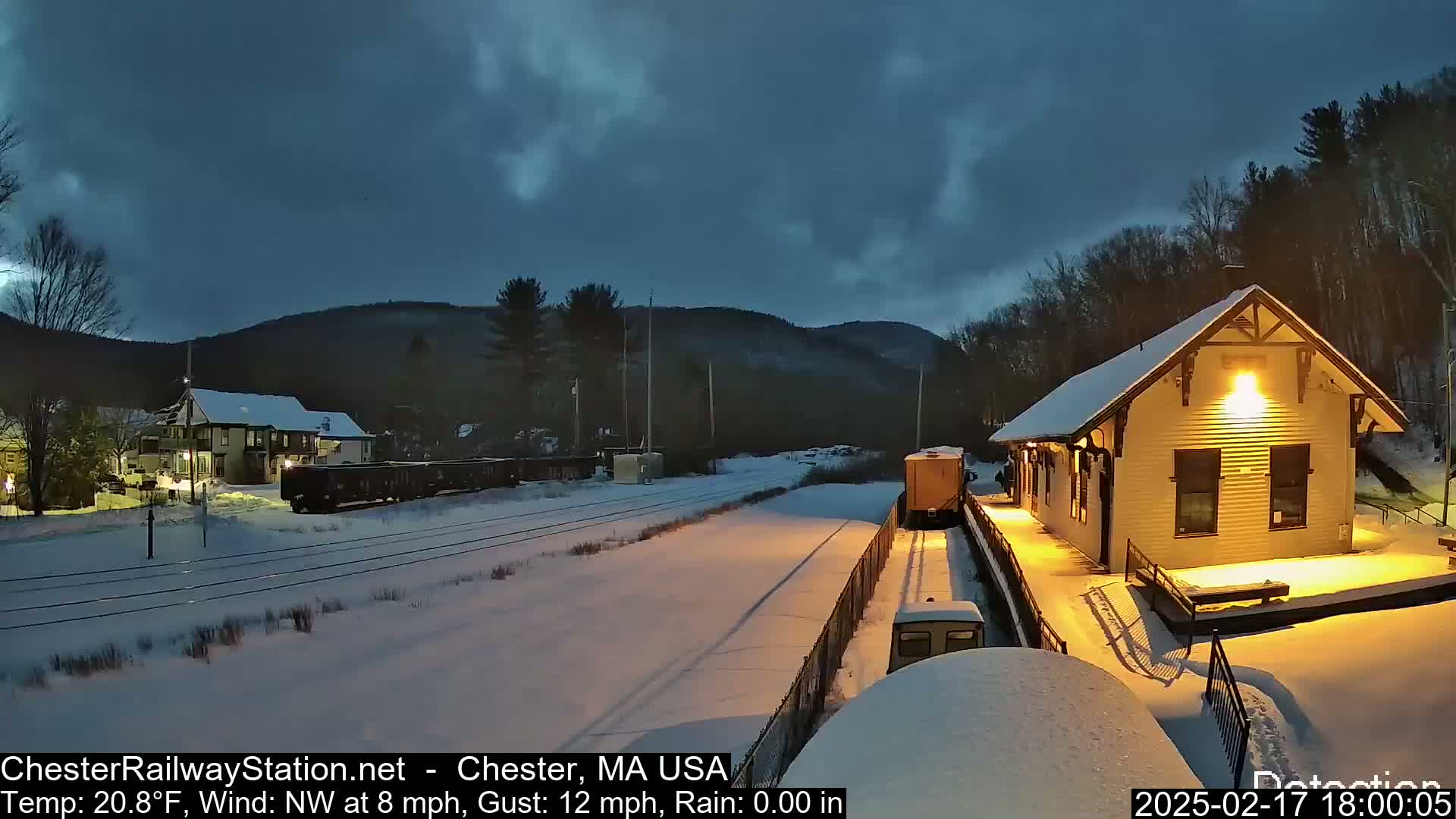 A snow-covered train station and railway line are illuminated at night under a dark, cloudy sky, with a train visible in the distance.