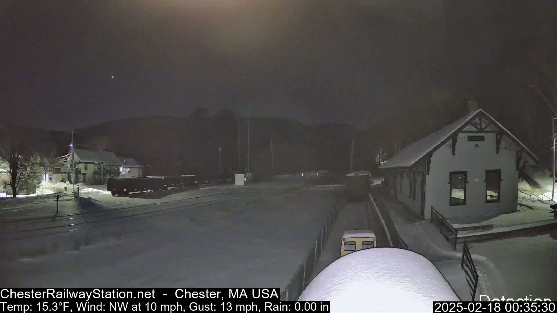 A snow-covered train station and surrounding area are visible at night under a dark sky, with a northwest wind at 10 mph and gusts up to 13 mph.