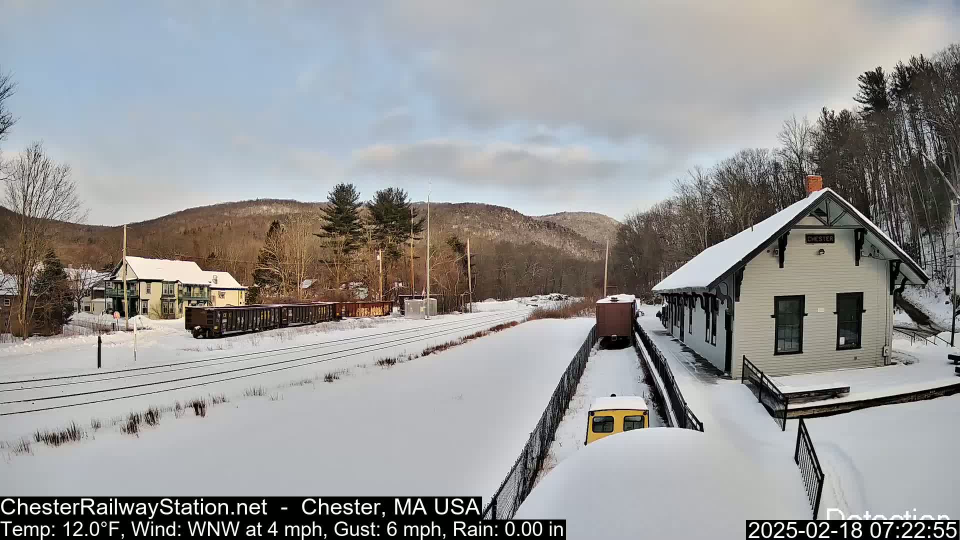 A snow-covered train station and rail yard are nestled in a winter landscape with a background of bare, snow-dusted hills under a partly cloudy sky.