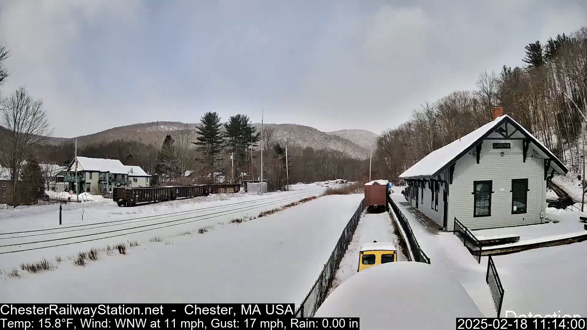 A snow-covered train station and rail yard are visible, with a train partially visible in the distance, under an overcast sky.
