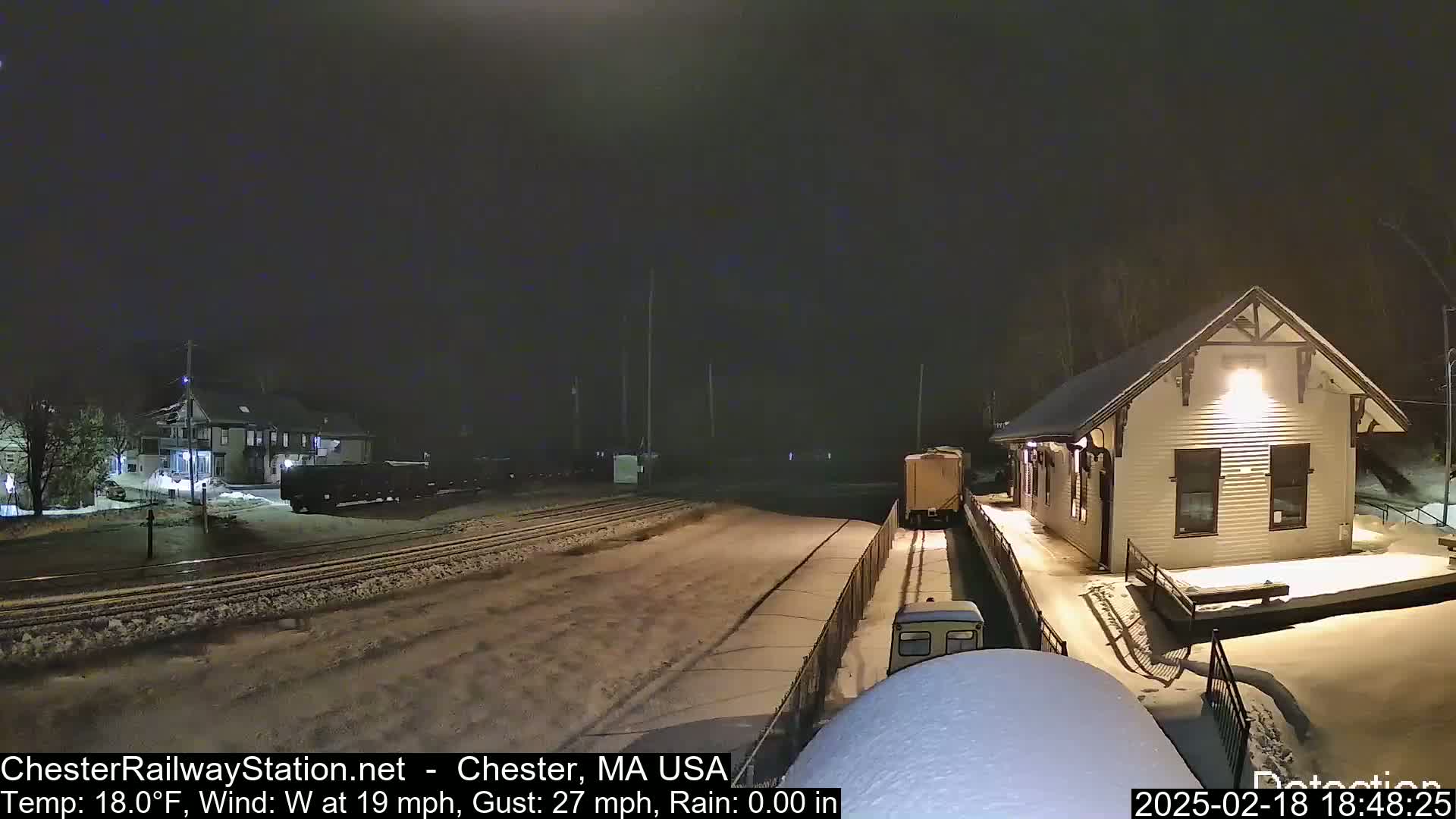 A snow-covered train station at night with a train passing by under windy, cold conditions.