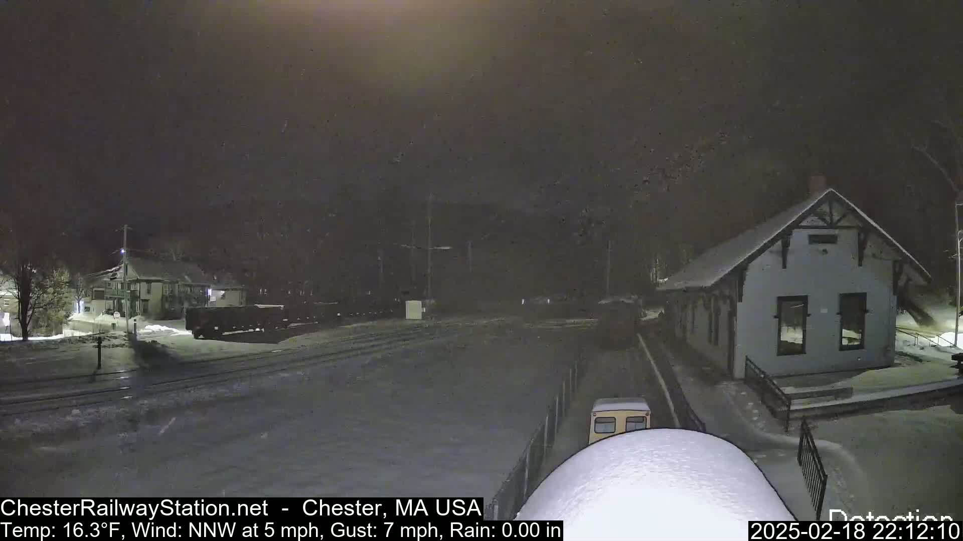 A snow-covered train station and railway yard are visible at night under rainy conditions.
