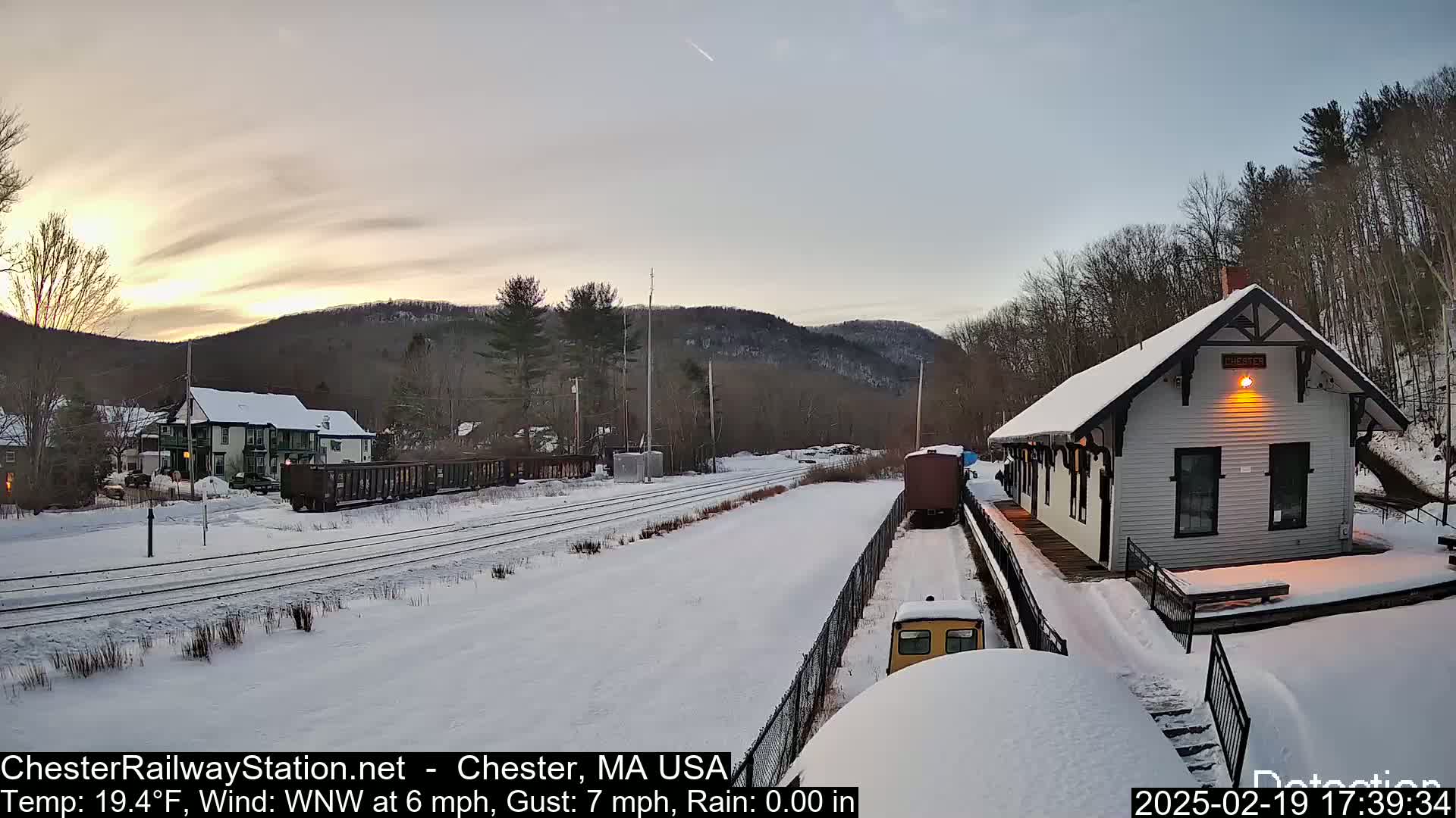 A snow-covered train station and rail yard are visible at sunset under a clear sky; a freight train is partially visible in the distance.