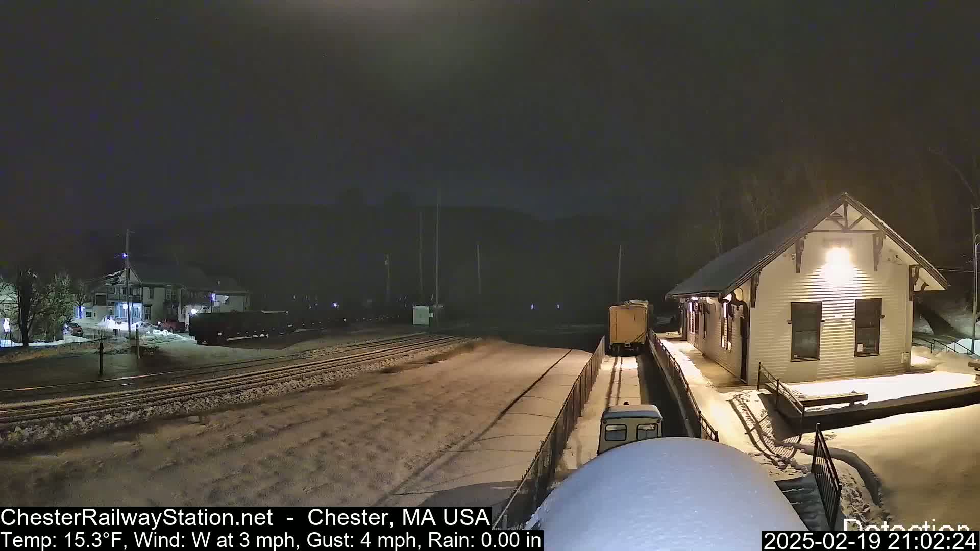 A snow-covered train station at night with a train passing by under a dark sky.