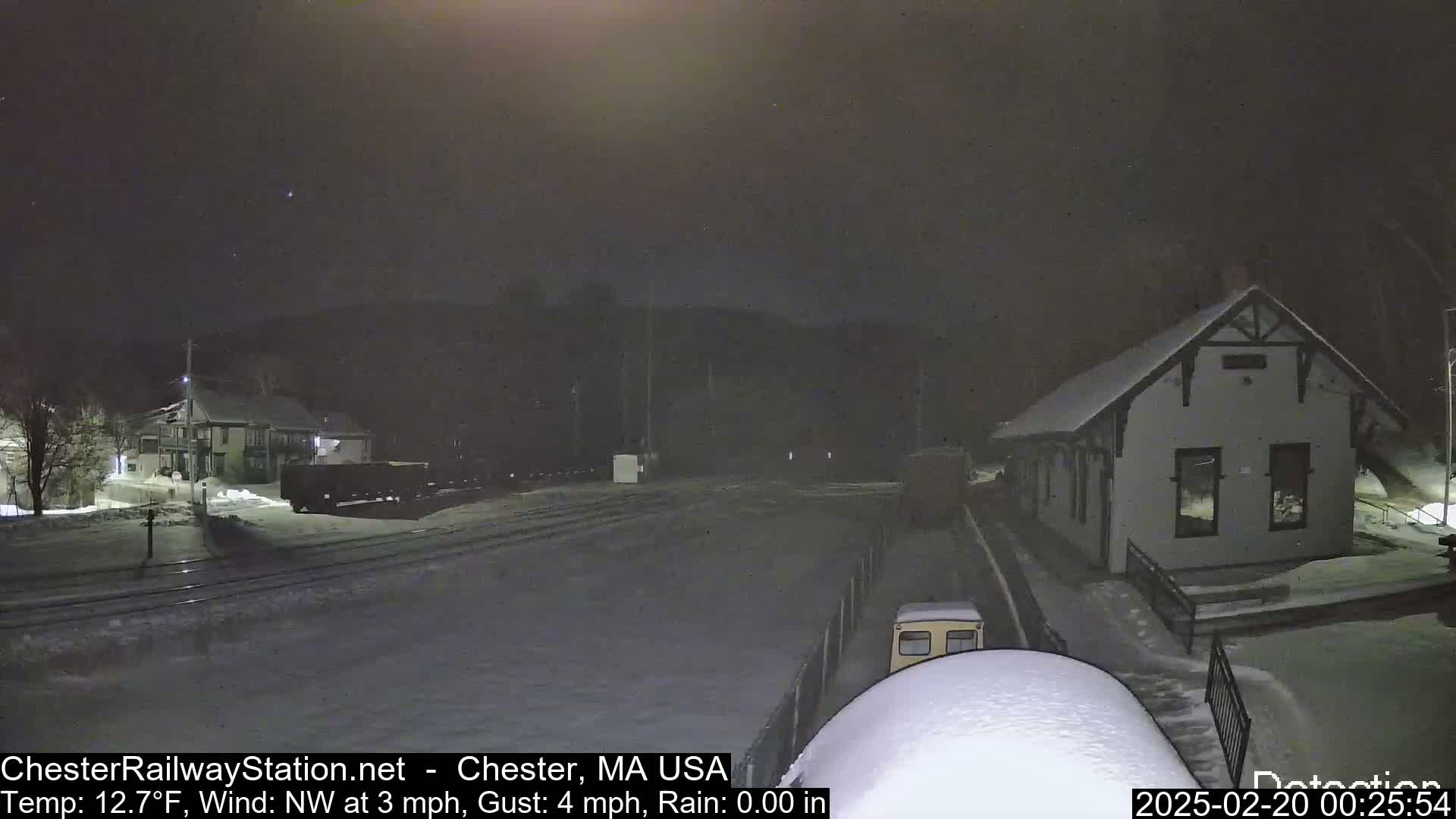 A snow-covered train station and railway yard are visible at night under light snowfall, with a train car parked nearby.