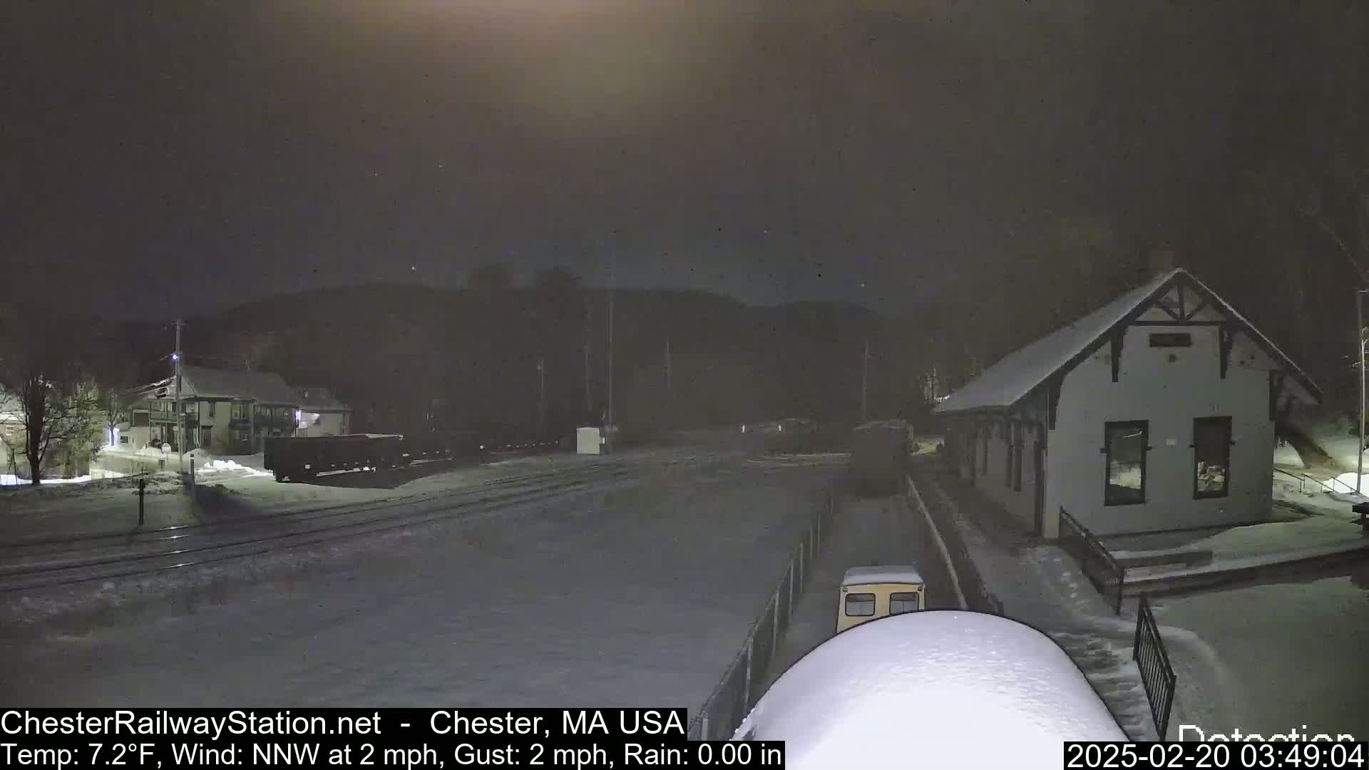 A snow-covered train station and railway line are visible at night under a dark sky, with light snow falling.