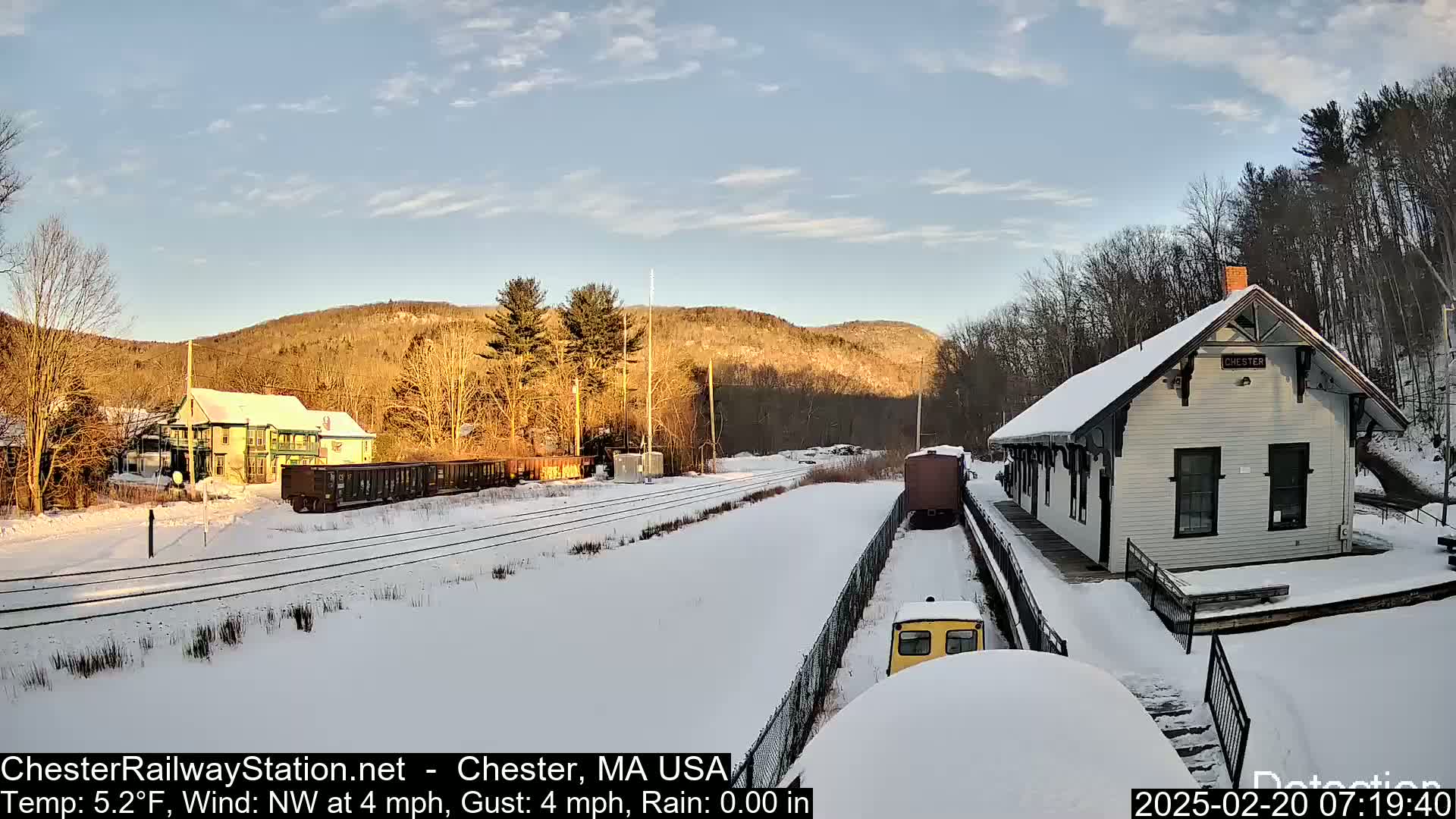 A snow-covered train station and railway line are visible, with a train and several railcars present under a clear, sunny sky.