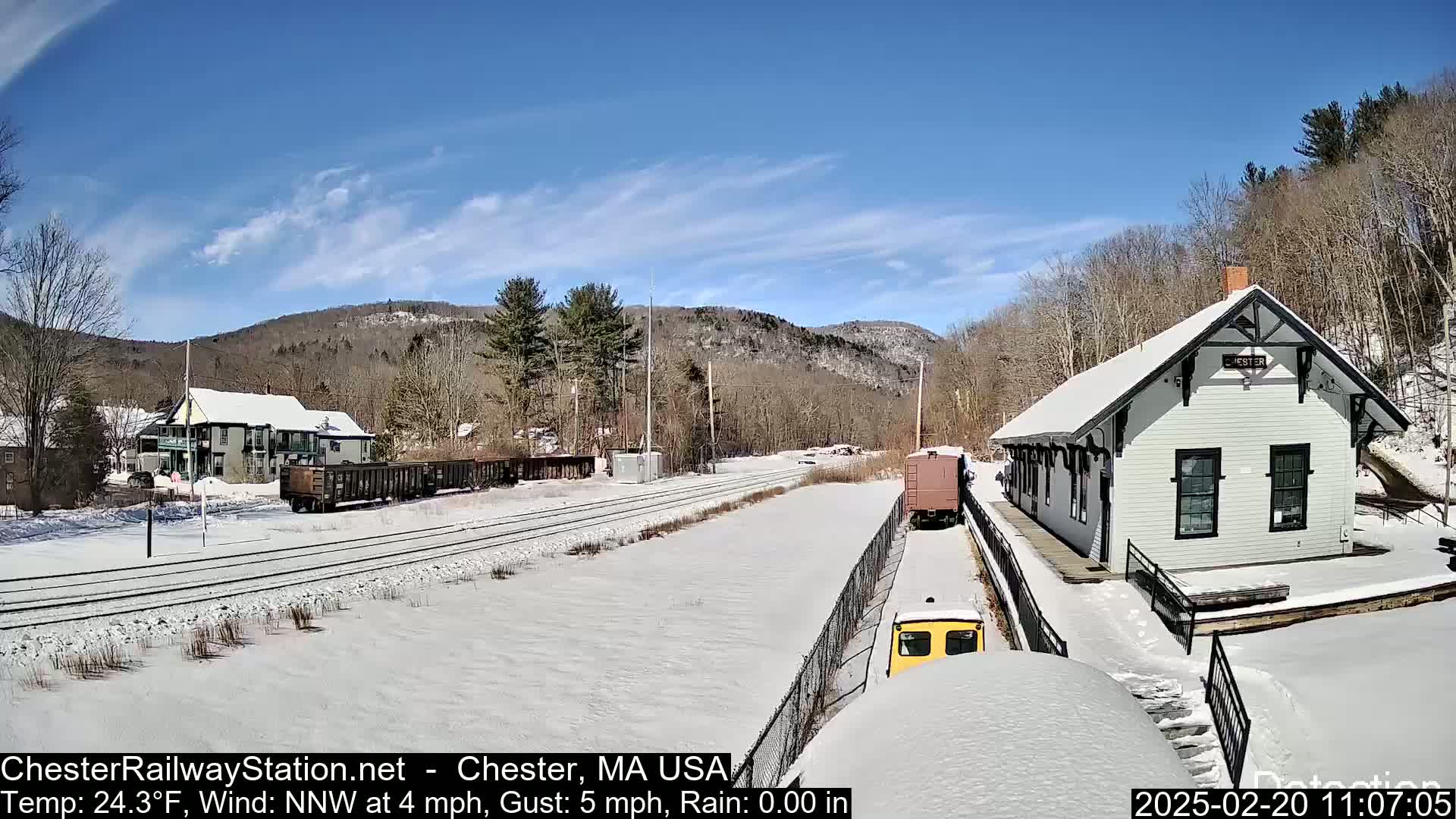 A snow-covered train station and tracks are situated in a valley amongst snow-covered hills and trees under a clear blue sky.