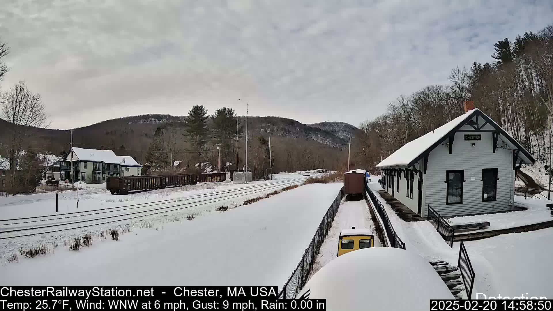 A snow-covered train station and railway line are situated in front of a snow-covered landscape with distant hills under an overcast sky.