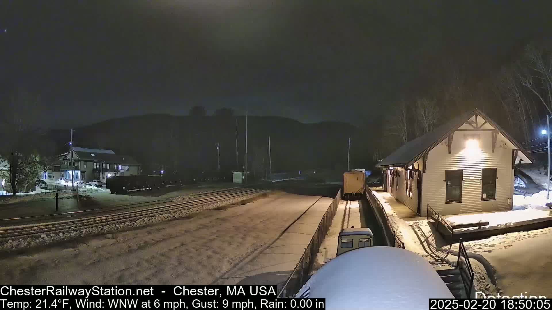 A snow-covered train station at night is illuminated, with a train partially visible in the foreground and another in the background under a dark sky.