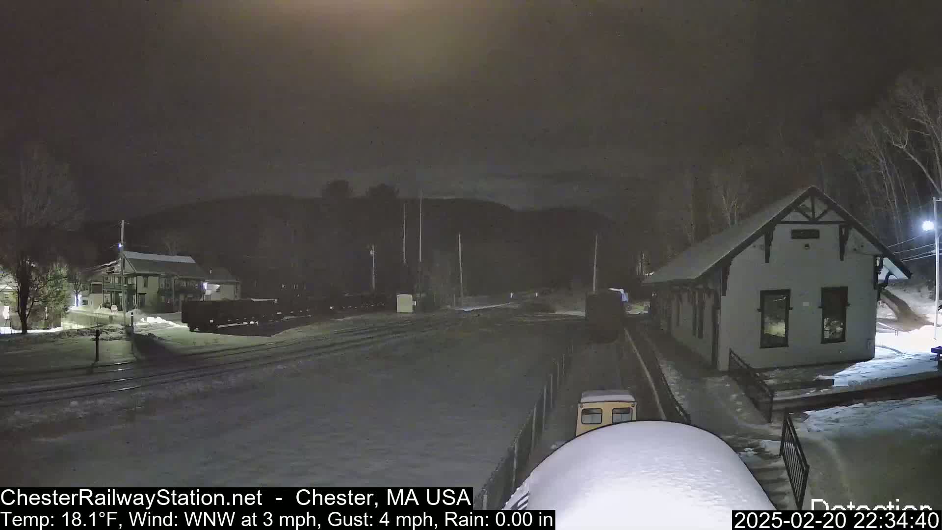 A nighttime view of a snow-covered train station and rail yard under a dark sky with a light wind and no rain.