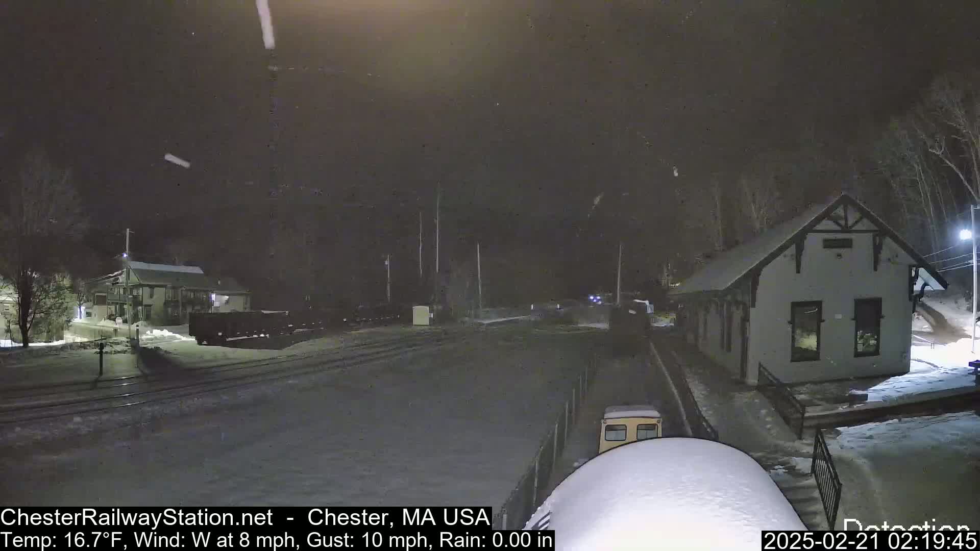 A nighttime snow scene shows a train car parked near a small building, with light snow falling.