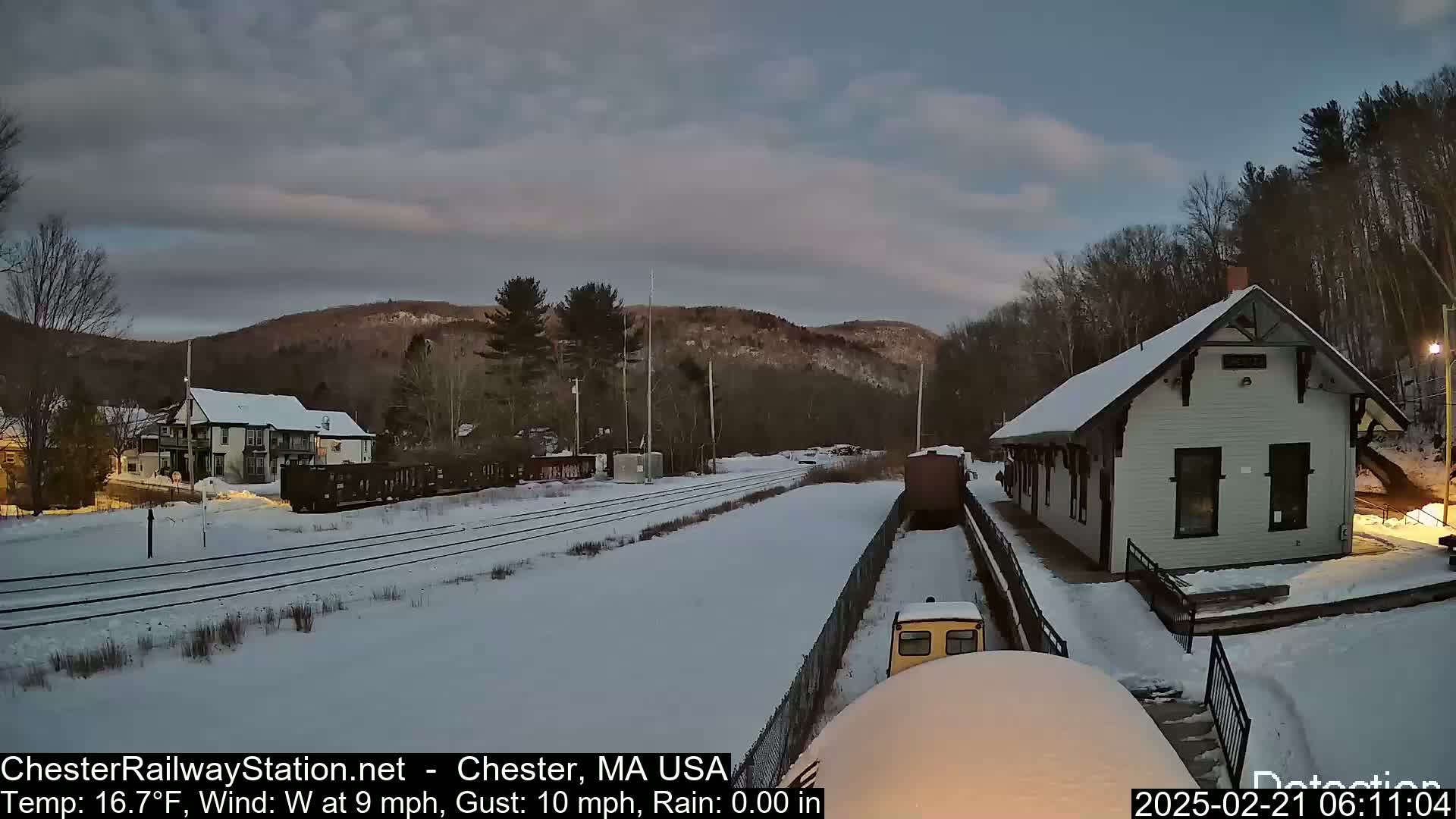 A snow-covered train station and tracks are visible under a cloudy, pre-dawn sky.