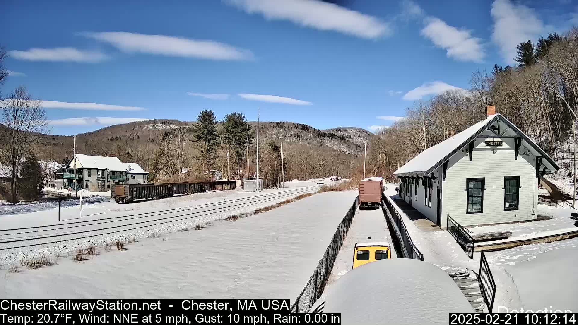 A snow-covered train station and rail yard are nestled in a valley under a partly sunny sky.