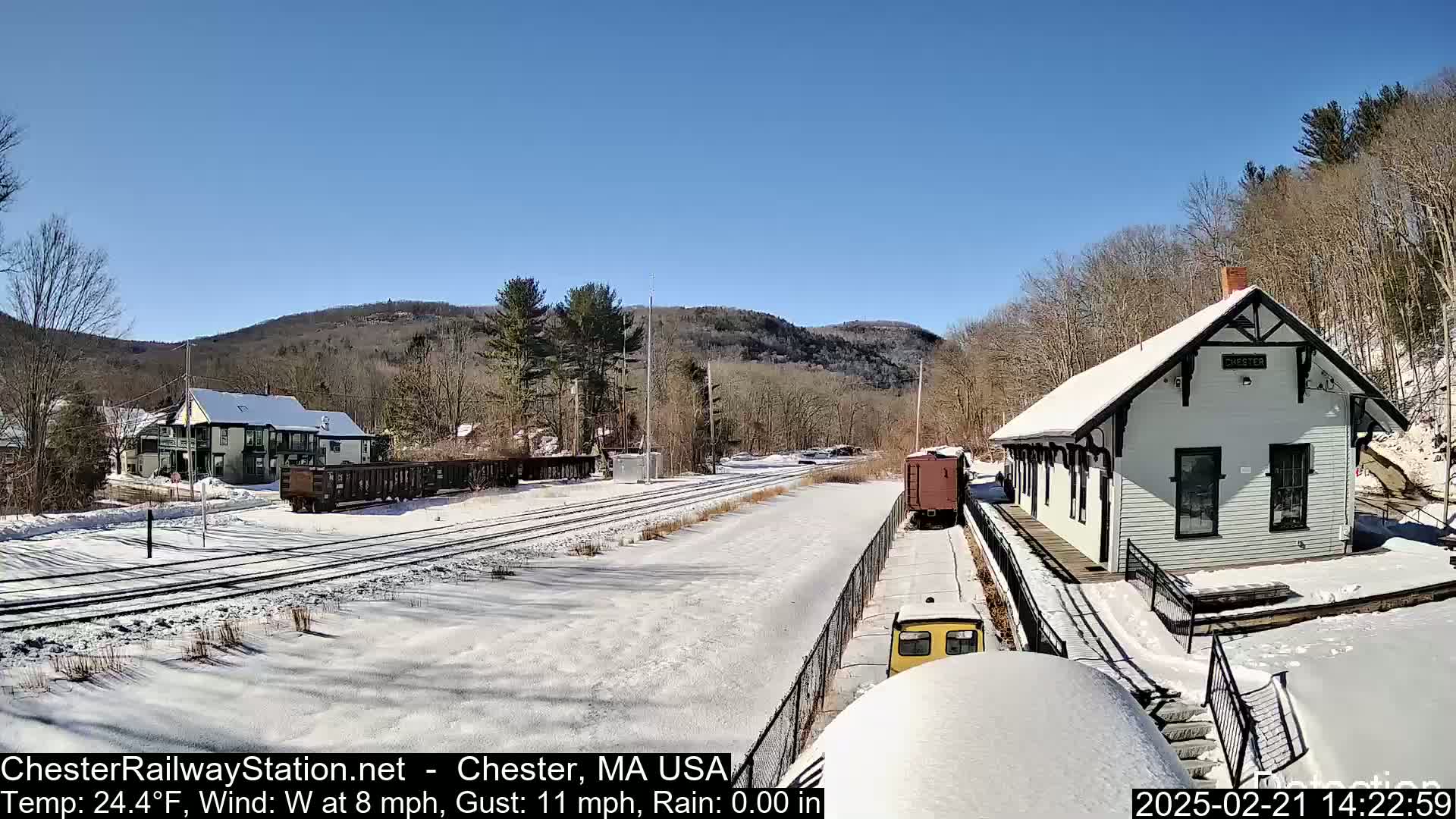 A snow-covered train station sits beside train tracks in a sunny, snowy landscape with a small town and hills in the background.