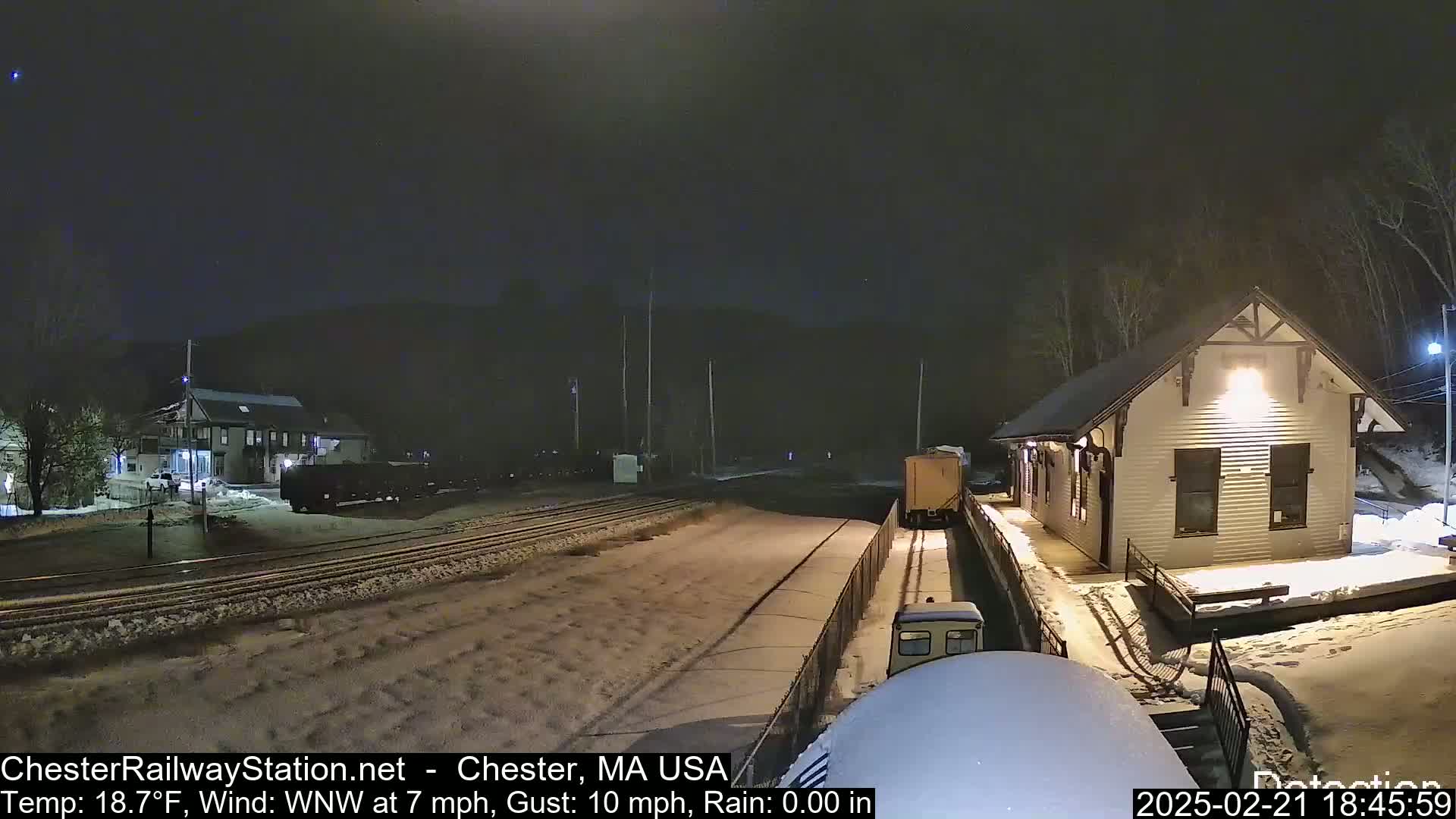 A snow-covered train station at night with a train approaching, under a dark sky with a wind blowing from the west-northwest at 7 miles per hour and gusts of 10 miles per hour.